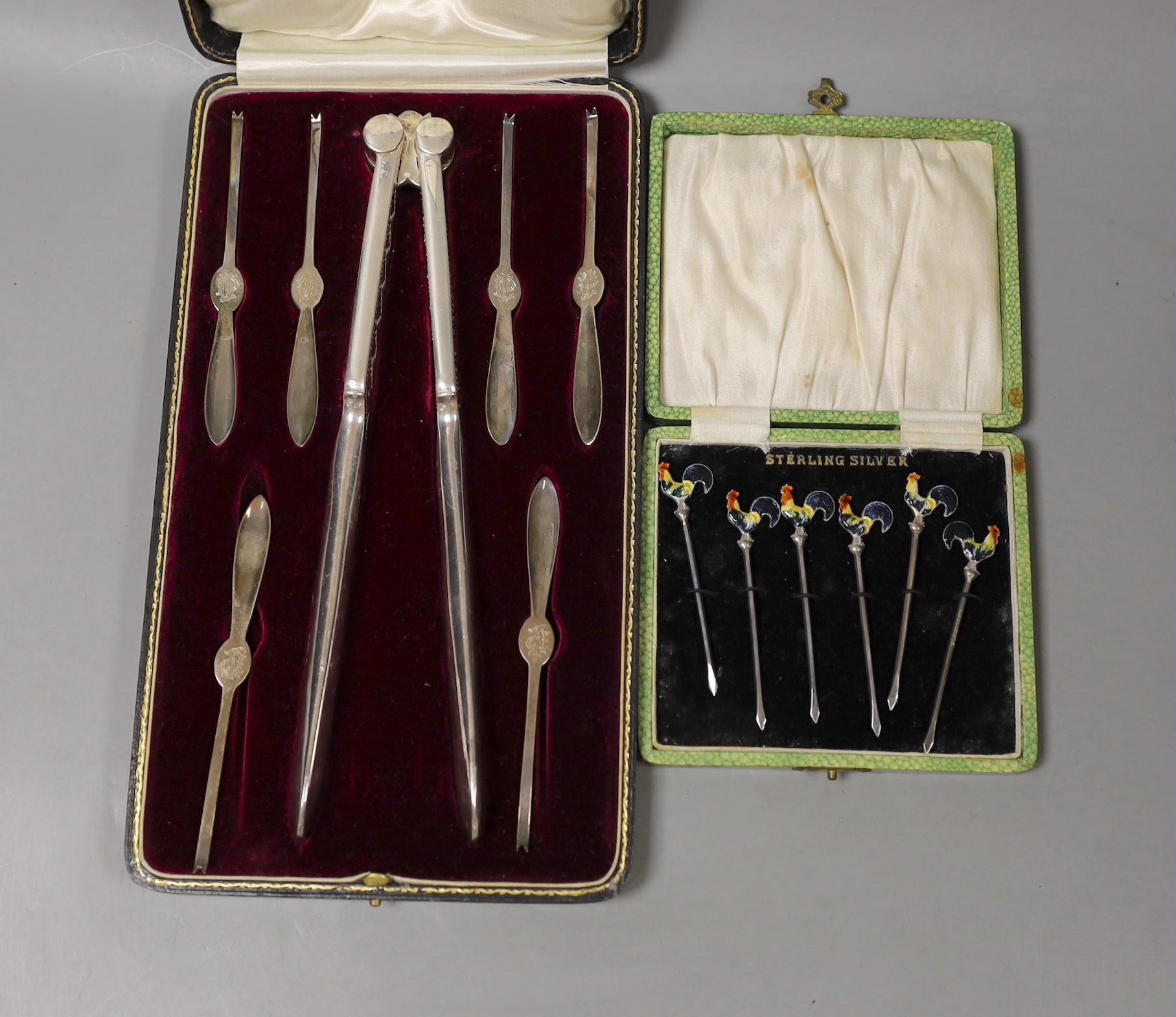 A George V cased set of six silver lobster picks and plated crackers and a similar set of silver and enamel cocktail sticks